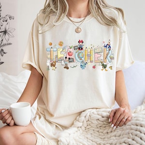 Peut inclure: T-shirt cr&egrave;me avec le mot "TEACHER" en lettres color&eacute;es de style dessin anim&eacute;. Les lettres sont d&eacute;cor&eacute;es de crayons, de livres, de pommes et de personnages Disney. Le t-shirt a des manches courtes.