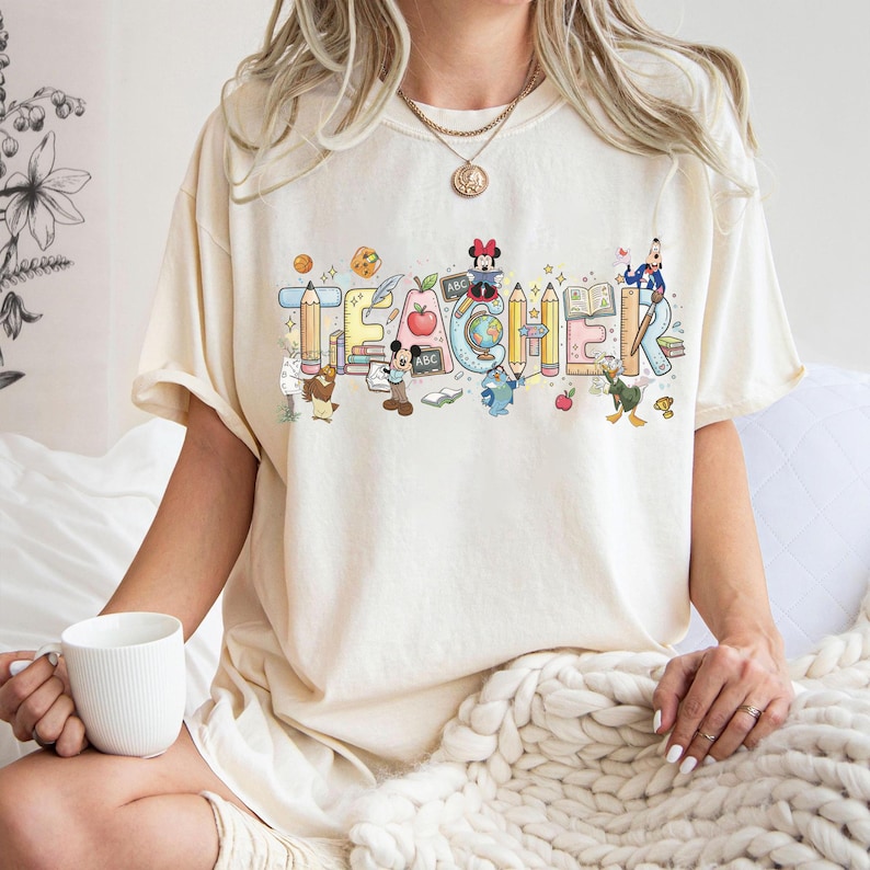 Peut inclure: T-shirt cr&egrave;me avec le mot "TEACHER" en lettres color&eacute;es de style dessin anim&eacute;. Les lettres sont d&eacute;cor&eacute;es de crayons, de livres, de pommes et de personnages Disney. Le t-shirt a des manches courtes.