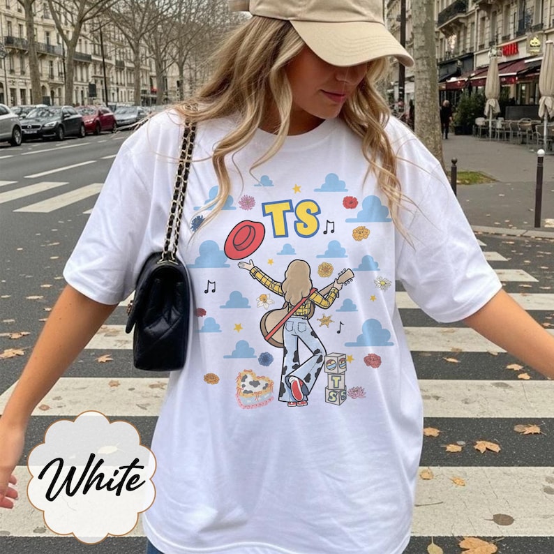 May include: A white t-shirt featuring a graphic print of a person with long hair playing a guitar, wearing a yellow plaid shirt and cow-print pants. The design includes blue clouds, a red hat, musical notes, and blocks with the letters TS.