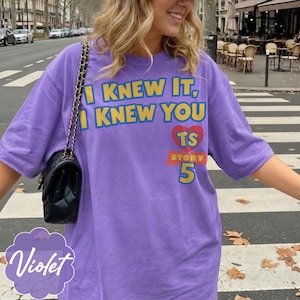 K&ouml;nnte beinhalten: Ein violettes kurz&auml;rmeliges T-Shirt mit dem gelben Text I KNEW IT, I KNEW YOU, einem roten Herz mit den Buchstaben TS, einem roten Banner mit dem Wort STORY und der gelben Zahl 5. Eine schwarze gesteppte Umh&auml;ngetasche mit Goldkette ist zu sehen.