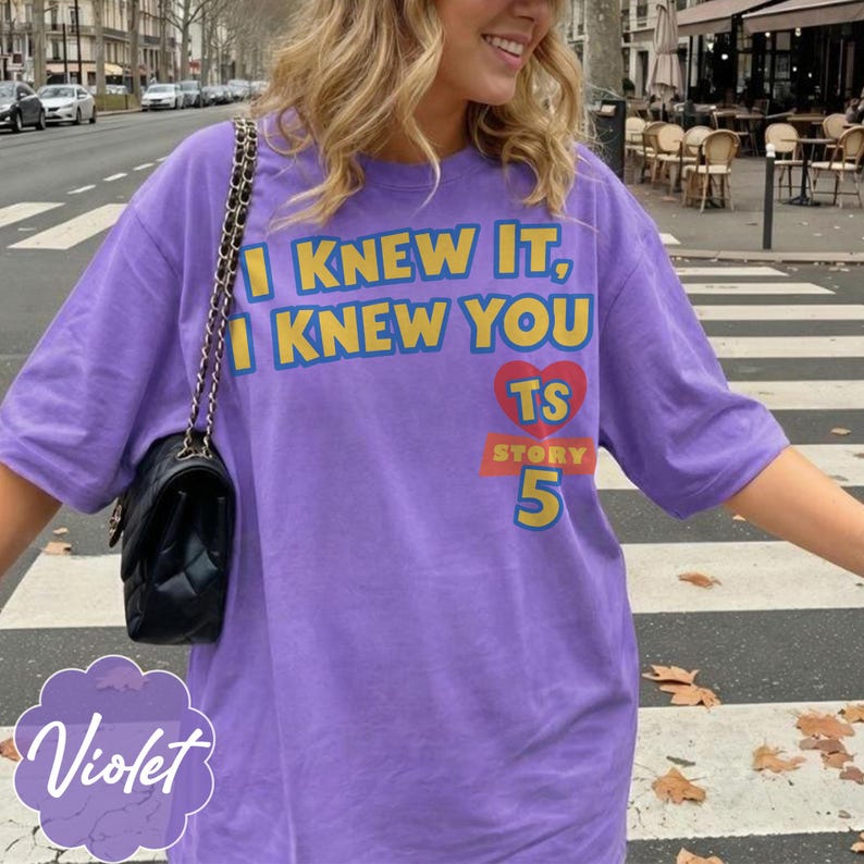 K&ouml;nnte beinhalten: Ein violettes kurz&auml;rmeliges T-Shirt mit dem gelben Text I KNEW IT, I KNEW YOU, einem roten Herz mit den Buchstaben TS, einem roten Banner mit dem Wort STORY und der gelben Zahl 5. Eine schwarze gesteppte Umh&auml;ngetasche mit Goldkette ist zu sehen.