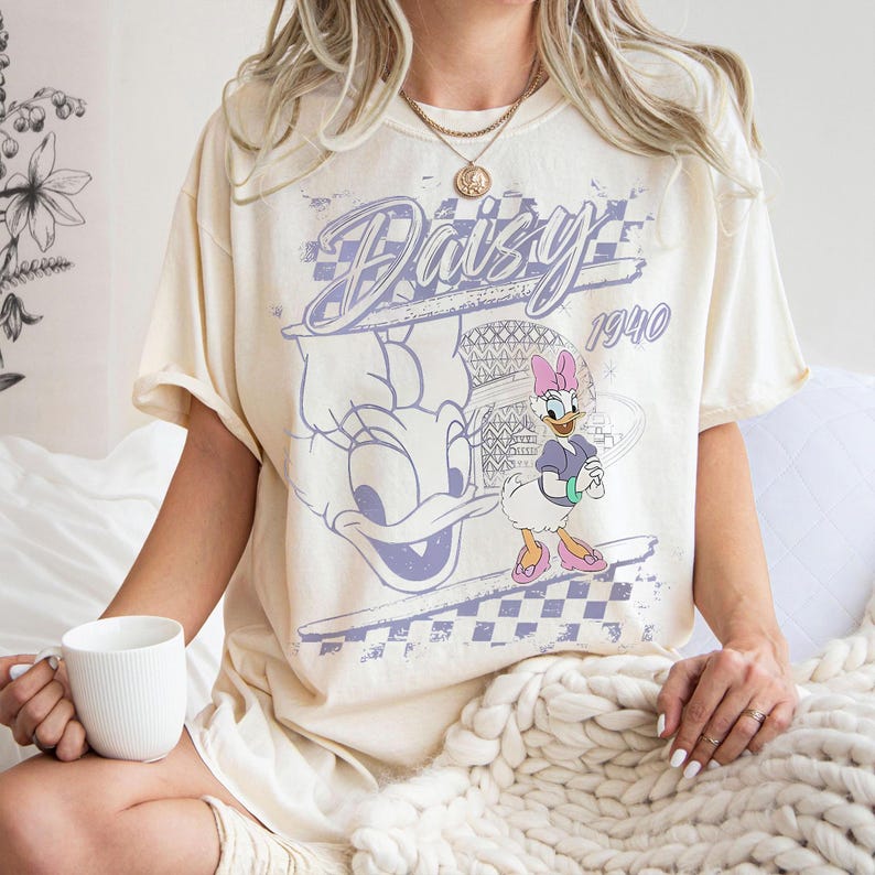 May include: Cream-colored t-shirt featuring a vintage-style graphic of Daisy Duck. The design includes the word "Daisy" in a stylized font, the year "1940", and an image of Daisy Duck. The shirt has short sleeves and a relaxed fit.