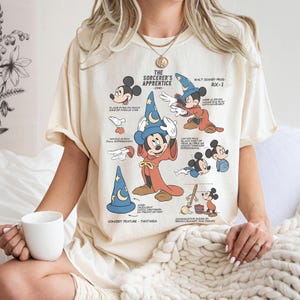May include: Cream-colored t-shirt featuring a graphic of Mickey Mouse in his Sorcerer's Apprentice costume from Fantasia. The design includes illustrations and text detailing the character's features and the film's title.