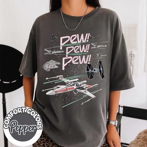 Könnte beinhalten: Dunkelgraues T-Shirt mit einem Weltraumschlacht-Design und dem Text "PEW! PEW! PEW!". Das Design zeigt Raumschiffe und Laserstrahlen in Pink und Grün. Das T-Shirt ist von Comfort Colors.