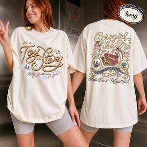 May include: An ivory t-shirt featuring a front graphic with rope-style text reading "it's a Toy Story Baby, just say yes" and a back graphic with rope-style text reading "Cowgirl like Me" and "Takes One to Know One" with a cowboy hat illustration.