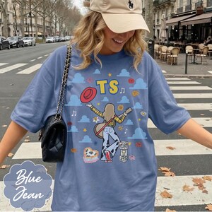 May include: A blue t-shirt featuring a graphic print of a person with a guitar, a red hat, clouds, stars, flowers, and blocks with the text TS. The design includes a heart-shaped cow print element and musical notes scattered across the fabric.