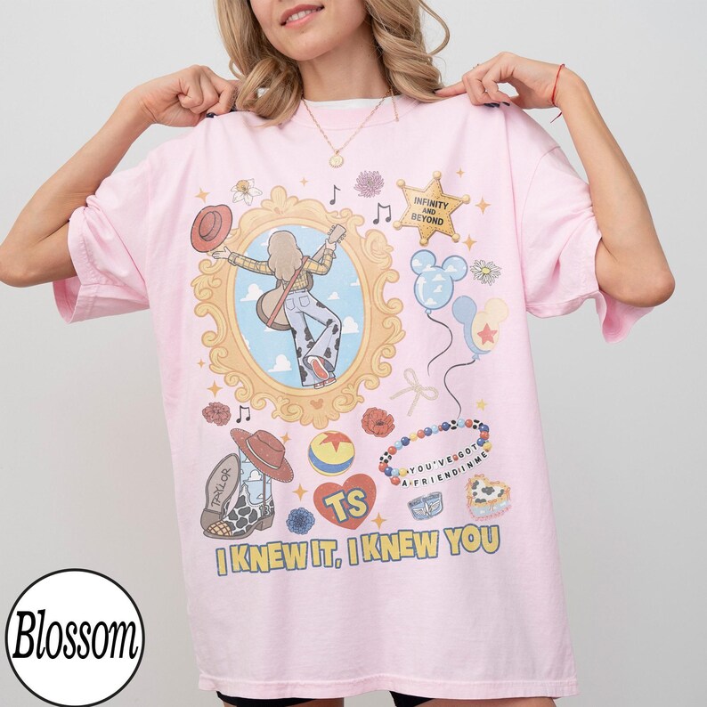 May include: A light pink t-shirt featuring a graphic print of a person with a guitar inside an ornate gold frame, surrounded by icons including a star with the text INFINITY AND BEYOND, a beaded bracelet with YOU'VE GOT A FRIEND IN ME, and text I KNEW IT, I KNEW YOU.
