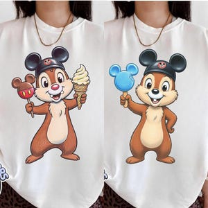 May include: White t-shirts featuring a cartoon chipmunk character wearing a black hat with a Mickey Mouse emblem. One chipmunk holds a red lollipop and an ice cream cone, the other holds a blue popsicle.