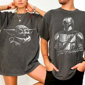 Pode incluir: Duas camisetas cinza escuro. Uma apresenta Grogu com o nome "Grogu" em escrita. A outra mostra The Mandalorian com o texto "The Mandalorian". Ambas as camisetas t&ecirc;m mangas curtas e um corte folgado.