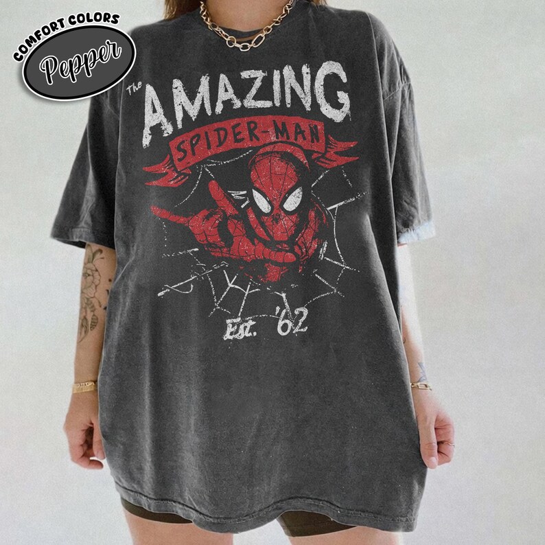 May include: A charcoal gray short-sleeve t-shirt featuring a distressed graphic of Spider-Man in red and black. The design includes the text The AMAZING SPIDER-MAN and Est. 62, set against a white spiderweb background with a red banner element.