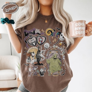 Op de afbeelding: Een bruin T-shirt met korte mouwen en een grafische print van personages en symbolen uit The Nightmare Before Christmas, waaronder Jack Skellington, Sally, Oogie Boogie en Zero, met een paarse strik en spinnenwebaccenten.