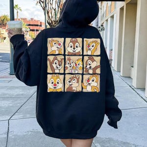 May include: A black hooded sweatshirt featuring a grid of nine cartoon chipmunk character portraits on the back. The chipmunks are in various poses and colors, set against a brown background. The person is holding a beverage in a clear cup.