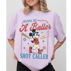 May include: Lilac-colored t-shirt featuring a vintage-style graphic of Mickey Mouse playing baseball. The design includes the text "WANNA BE A Baller SHOT CALLER" in red and blue. The shirt is a casual, unisex style.