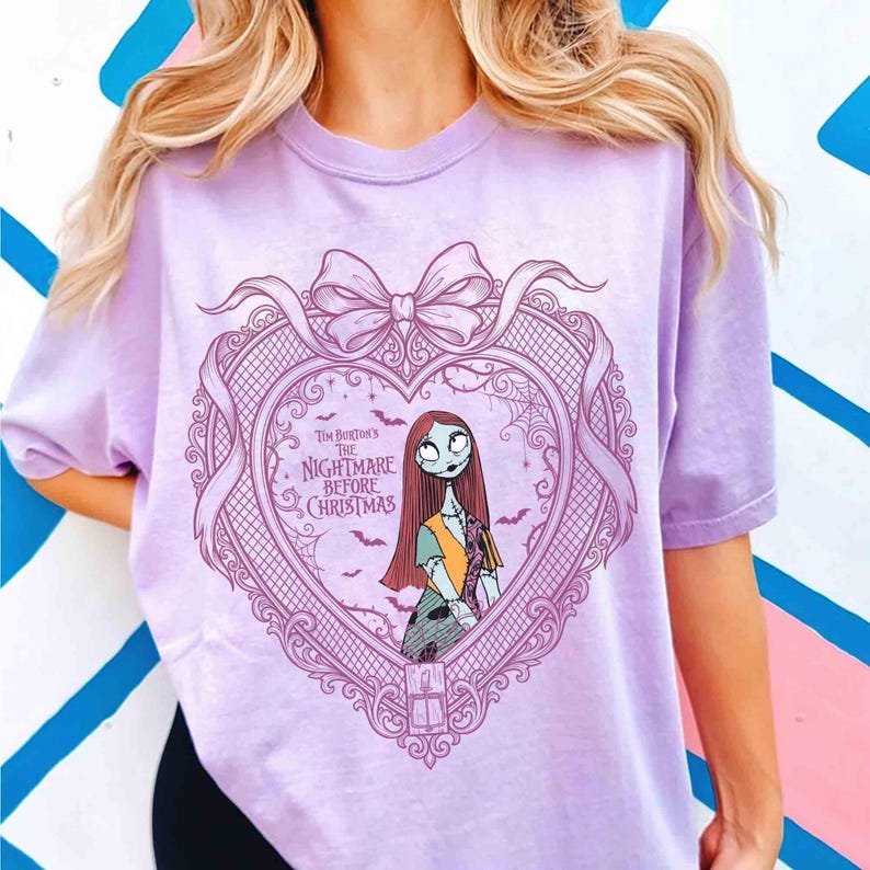 May include: A light purple t-shirt featuring a heart-shaped design with a bow at the top. The design includes an illustration of Sally from Tim Burton's "The Nightmare Before Christmas" and the movie title.