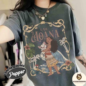 May include: Dark gray t-shirt featuring a graphic of Moana holding a pig, with the text "I AM MOANA." The design includes palm trees, flowers, and a rooster. The shirt has the words "Comfort Colors" and "Pepper" printed on it.