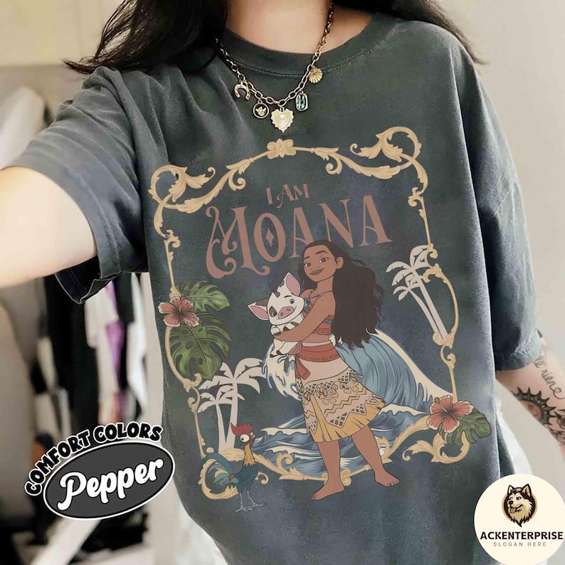 May include: Dark gray t-shirt featuring a graphic of Moana holding a pig, with the text "I AM MOANA." The design includes palm trees, flowers, and a rooster. The shirt has the words "Comfort Colors" and "Pepper" printed on it.