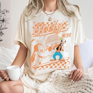 May include: Cream-colored t-shirt featuring a graphic of Goofy in orange and black. The design includes the word "Goofy" in a stylized font, the year "1938", and a checkered pattern. The shirt has short sleeves.
