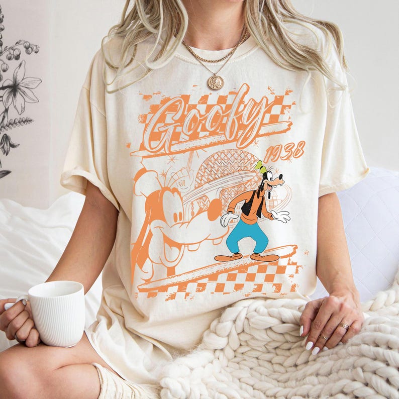 May include: Cream-colored t-shirt featuring a graphic of Goofy in orange and black. The design includes the word "Goofy" in a stylized font, the year "1938", and a checkered pattern. The shirt has short sleeves.