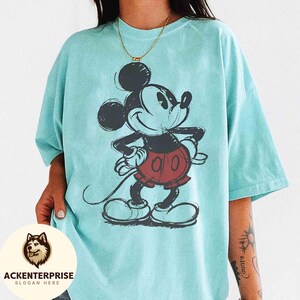 Puede incluir: Camiseta azul claro con un dibujo de Mickey Mouse en negro, rojo y blanco. El personaje de dibujos animados sonr&iacute;e con las manos en las caderas. La camiseta tiene mangas cortas y un corte holgado.