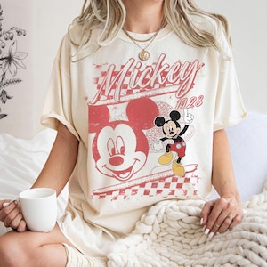 May include: Cream-colored t-shirt featuring a distressed red graphic of Mickey Mouse and the text "Mickey 1928". The design includes a large Mickey face and a smaller full-body illustration. The shirt has short sleeves.