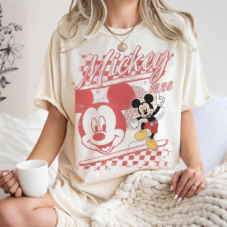 May include: Cream-colored t-shirt featuring a distressed red graphic of Mickey Mouse and the text "Mickey 1928". The design includes a large Mickey face and a smaller full-body illustration. The shirt has short sleeves.