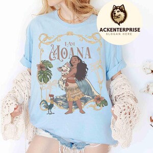 May include: Light blue t-shirt featuring a graphic of Moana holding a pig, with the text "I AM MOANA" in gold lettering. The design includes tropical flowers and a rooster. The shirt has rolled-up sleeves.