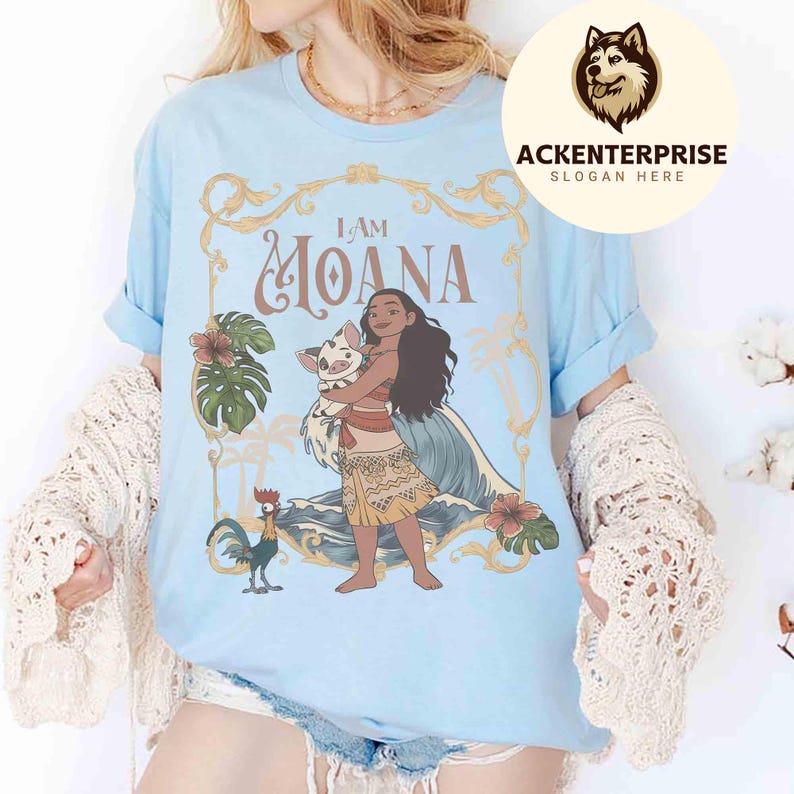 May include: Light blue t-shirt featuring a graphic of Moana holding a pig, with the text "I AM MOANA" in gold lettering. The design includes tropical flowers and a rooster. The shirt has rolled-up sleeves.
