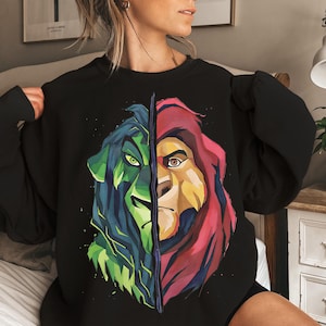May include: Black sweatshirt featuring a split-face design of a lion. One side is green and blue, the other is red and yellow. The graphic is a stylized illustration of a lion's face, with detailed eyes and fur.