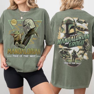 May include: Olive green t-shirt featuring a graphic of The Mandalorian and Grogu. The front design includes the text "MANDALORIAN" and "THIS IS THE WAY". The back of the shirt displays the Star Wars logo and the text "THE MANDALORIAN AND GROGU".