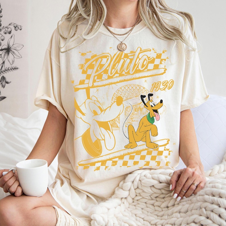 May include: Cream-colored t-shirt featuring a yellow graphic of Pluto, the Disney cartoon dog, with the text "Pluto 1930". The design includes a checkered pattern and a cartoon illustration of Pluto.