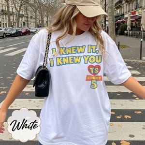 K&ouml;nnte beinhalten: Ein wei&szlig;es kurz&auml;rmeliges T-Shirt mit dem blauen Schriftzug I KNEW IT, I KNEW YOU &uuml;ber einem roten Herz mit den Buchstaben TS, wobei das Wort STORY und die Zahl 5 darunter in einem gelb-roten Blockdesign auf der Vorderseite aufgedruckt sind.
