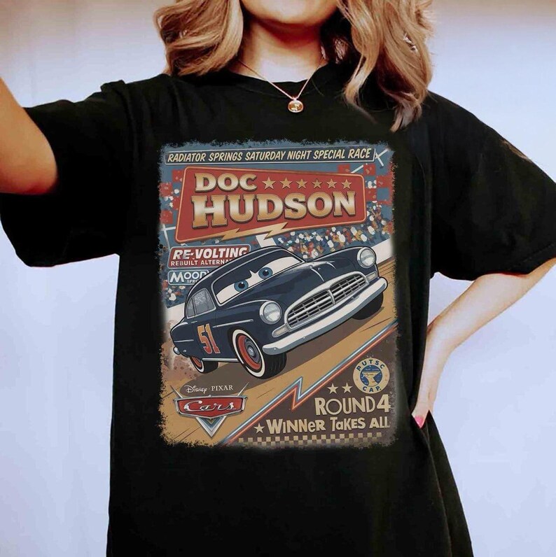 May include: Black t-shirt featuring a graphic of Doc Hudson from the Disney Pixar Cars movie. The design includes the text "RADIATOR SPRINGS SATURDAY NIGHT SPECIAL RACE" and "ROUND 4 WINNER TAKES ALL". The car is navy blue with the number 51.