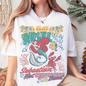 May include: White t-shirt featuring a colorful graphic of Ariel the mermaid, Flounder, and Sebastian from The Little Mermaid. The design includes the text "Ariel Ocean Tour 1991" and "Sebastian with the Hot Crustacean Band."