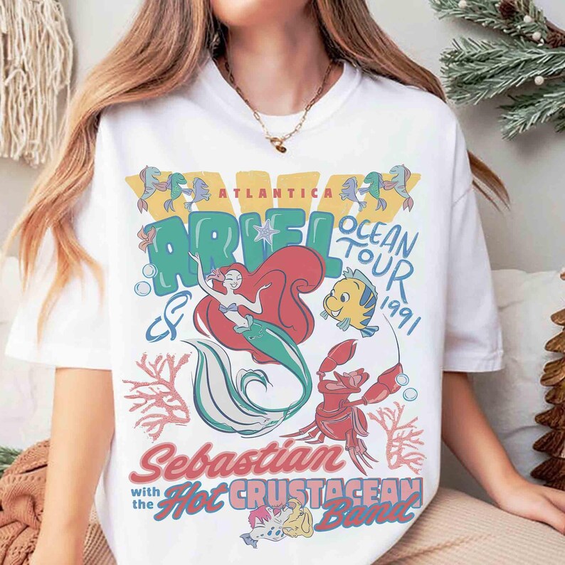 May include: White t-shirt featuring a colorful graphic of Ariel the mermaid, Flounder, and Sebastian from The Little Mermaid. The design includes the text "Ariel Ocean Tour 1991" and "Sebastian with the Hot Crustacean Band."