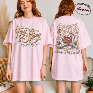 May include: A light pink short-sleeve t-shirt featuring a front graphic with rope-style text reading "it's a Toy Story Baby, just say "yes"" and a back graphic with rope borders, a hat, and text reading "Cowgirl like Me Takes One to Know One."