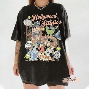 Może przedstawiać: Czarny t-shirt z kolorową grafiką z napisem "Hollywood Studios" i różnymi postaciami z kreskówek. Projekt zawiera statek kosmiczny z Gwiezdnych Wojen, aparat fotograficzny, dinozaura i inne animowane postacie. Widoczne jest również słowo "Fantasmic".