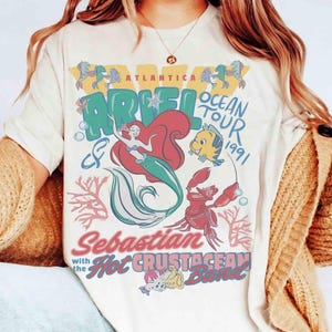 May include: Cream-colored t-shirt featuring a colorful graphic of Ariel from The Little Mermaid. The design includes Ariel, Flounder, Sebastian, and the text "Atlantica Ariel Ocean Tour 1991" and "Sebastian with the Hot Crustacean Band."