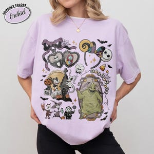 Op de afbeelding: Een lichtpaars t-shirt met een grafische print van personages en motieven uit The Nightmare Before Christmas, waaronder Jack Skellington, Sally, Oogie Boogie, Zero en diverse griezelige elementen zoals vleermuizen, spinnenwebben en snoep.