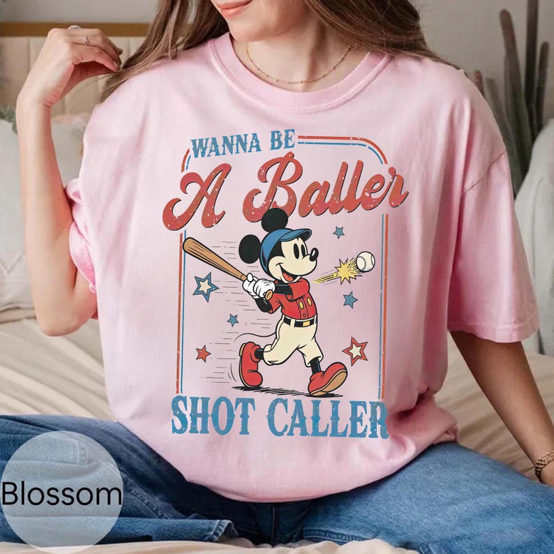 May include: Pink t-shirt featuring a graphic of Mickey Mouse swinging a baseball bat. The text reads "Wanna Be A Baller Shot Caller" in a vintage style. The shirt has short sleeves and a relaxed fit.