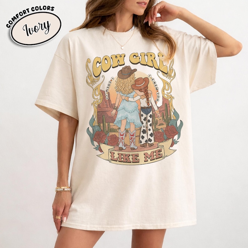May include: An ivory short-sleeve t-shirt featuring a graphic print of two figures in cowboy hats standing in a desert landscape with cacti and roses. The text "COW GIRL" appears above the figures and "LIKE ME" is printed on a banner below.