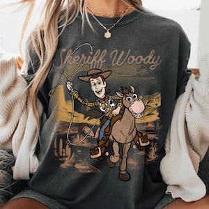 May include: Dark gray sweatshirt featuring Sheriff Woody from Toy Story. The graphic shows Woody riding a horse with a lasso, with the words "Sheriff Woody" above. The design has a vintage, distressed look.