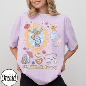 May include: A light purple t-shirt featuring a graphic print with a central gold ornate frame containing a person with a guitar, a cowboy hat, a star badge with the text INFINITY AND BEYOND, balloons, a beaded bracelet, and the text I KNEW IT, I KNEW YOU.
