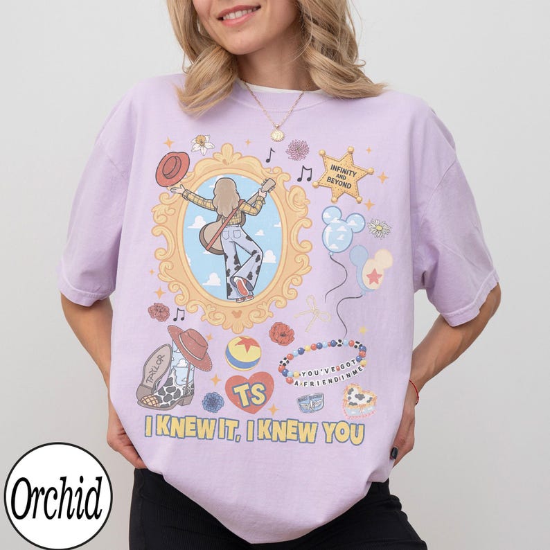 May include: A light purple t-shirt featuring a graphic print with a central gold ornate frame containing a person with a guitar, a cowboy hat, a star badge with the text INFINITY AND BEYOND, balloons, a beaded bracelet, and the text I KNEW IT, I KNEW YOU.