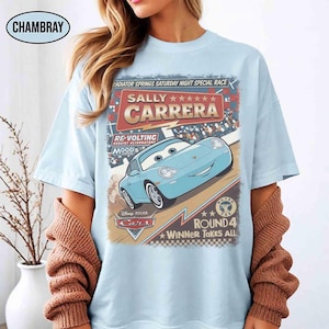 May include: Light blue t-shirt featuring a graphic of Sally Carrera from the Disney Pixar Cars movie. The design includes text that reads "Radiator Springs Saturday Night Special Race" and "Round 4 Winner Takes All."
