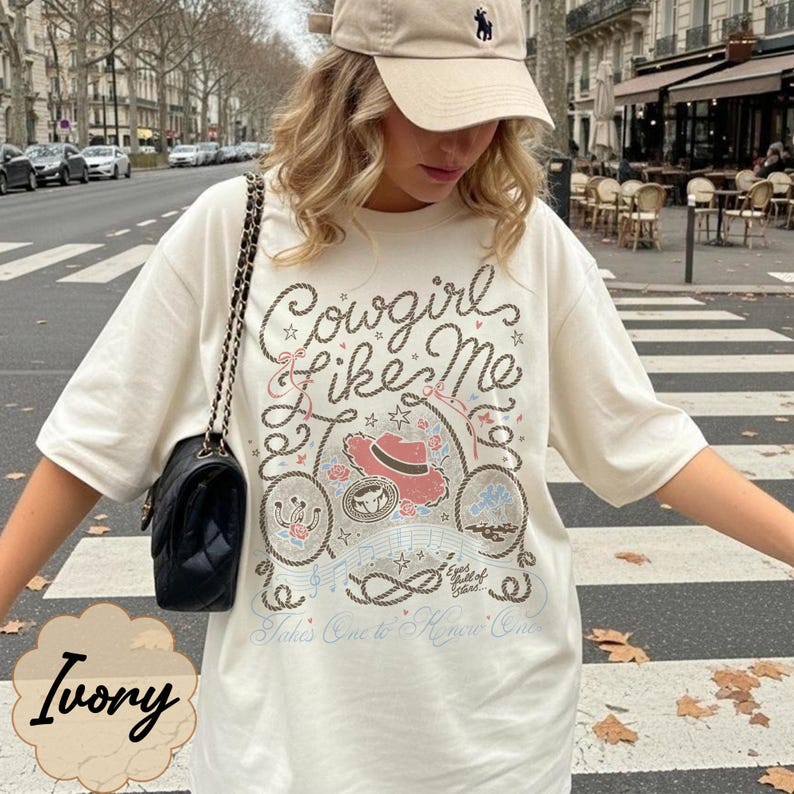 Puede incluir: Una camiseta de manga corta color marfil con un estampado gr&aacute;fico que incluye el texto Cowgirl like Me, un sombrero de vaquero rojo, una herradura y motivos de cuerda. El dise&ntilde;o incluye notas musicales y el texto Eyes full of stars y Takes One to Know One.