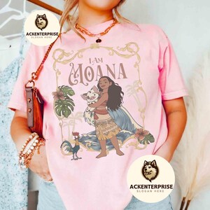 May include: Pink t-shirt featuring the words "I AM MOANA" in a decorative frame, with a cartoon illustration of Moana holding a pig. The design includes tropical flowers, a rooster, and ocean waves. The shirt has the logo "ACKENTERPRISE" on it.