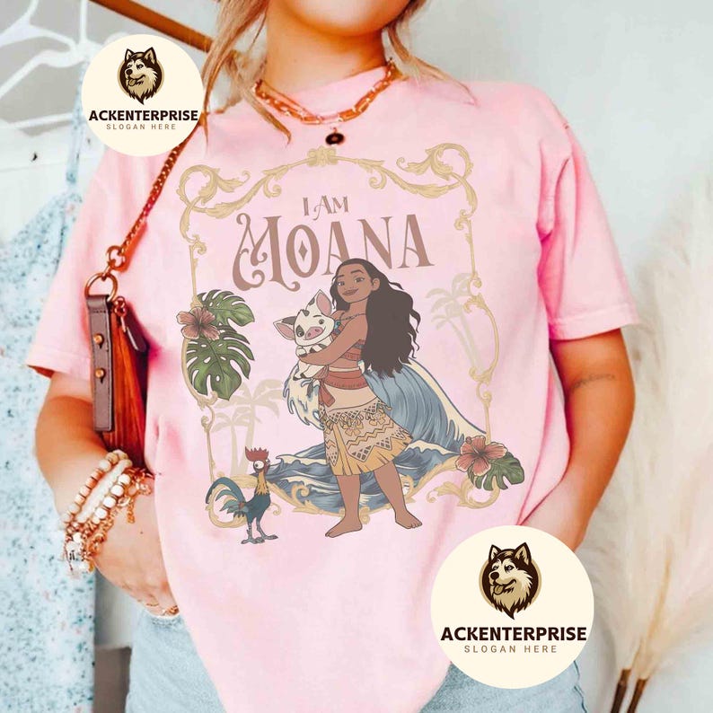 May include: Pink t-shirt featuring the words "I AM MOANA" in a decorative frame, with a cartoon illustration of Moana holding a pig. The design includes tropical flowers, a rooster, and ocean waves. The shirt has the logo "ACKENTERPRISE" on it.