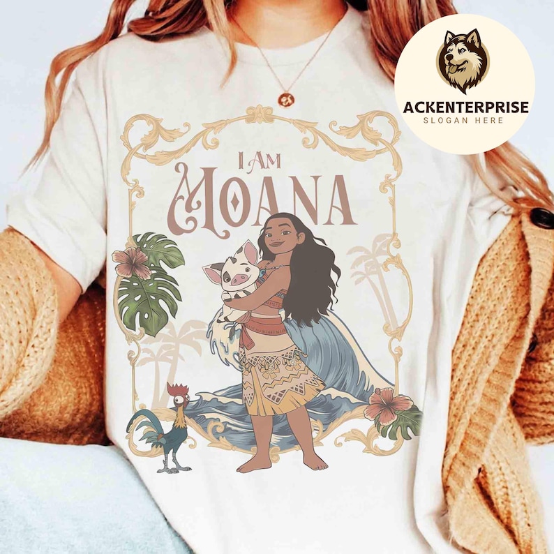 May include: Cream-colored t-shirt featuring a graphic of Moana holding a pig, with the text "I AM MOANA." The design includes tropical elements like flowers, palm trees, and a rooster. The shirt has a round neckline and short sleeves.
