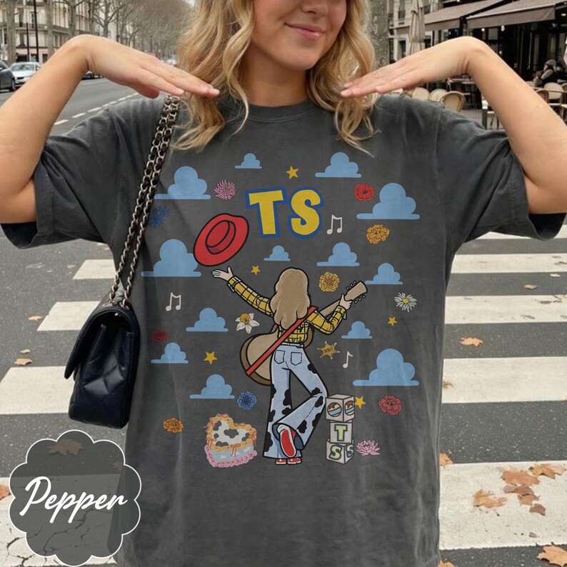 May include: A charcoal gray short-sleeve t-shirt featuring a graphic print of a person with blonde hair wearing a yellow plaid shirt and cow-print pants, holding a guitar. The design includes a red hat, blue clouds, stars, flowers, and blocks with the text TS.