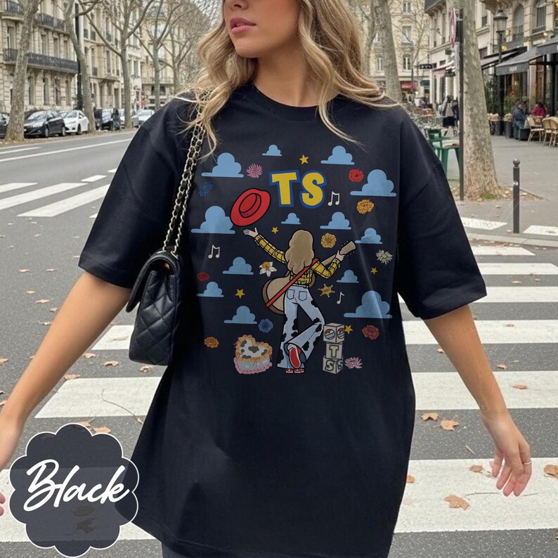 May include: A black short-sleeve t-shirt featuring a graphic print of a person with long hair playing a guitar, surrounded by blue clouds, red hats, flowers, musical notes, and the letters TS. The design includes stacked blocks with TS.